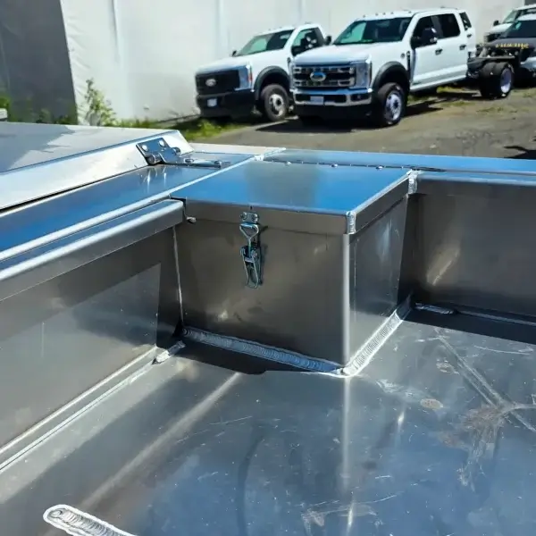 A close-up of the Lynx 7 metal storage box with latch, designed for wildfire equipment and placed in a shiny aluminum truck bed. In the background, two white pickups are parked outdoors near a white building.