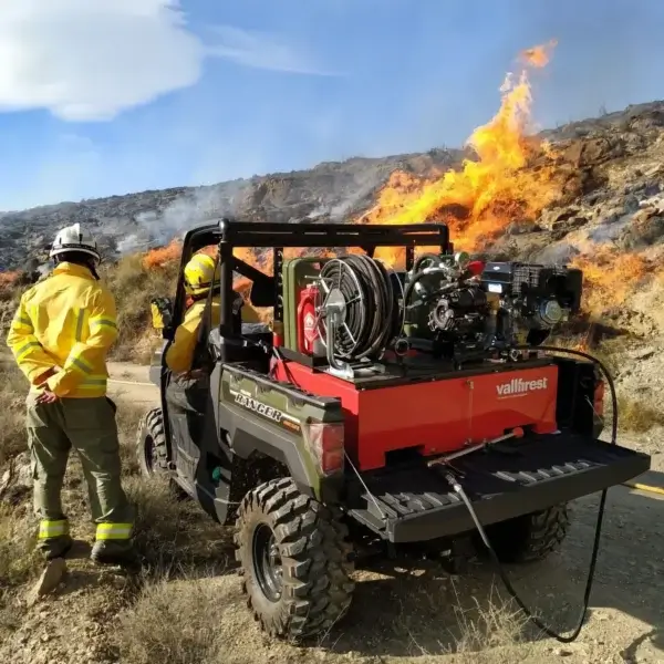 Two firefighters in yellow gear stand by a UTV Skid Unit, equipped for wildfires as used in Canada, observing a wildfire burning on a rocky hillside beneath a partly cloudy sky.