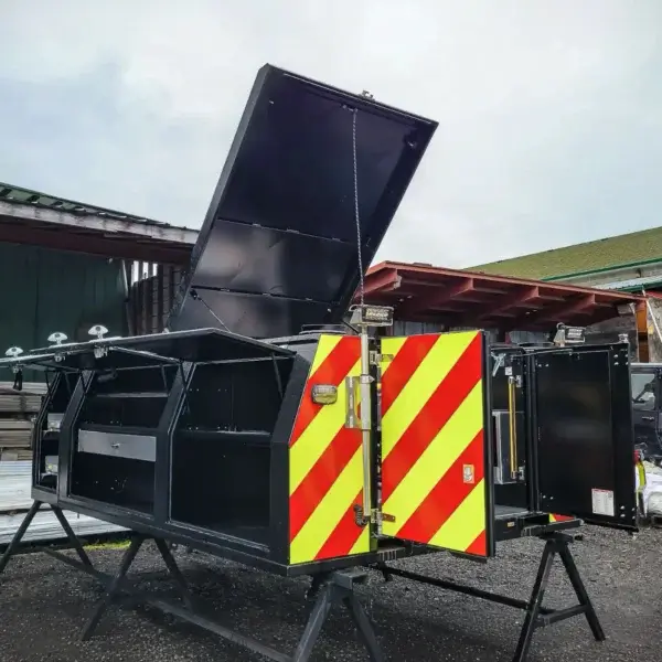 The Wildcat 1150, a large black firefighter utility storage box with red and yellow reflective stripes, is open to reveal compartments. It sits on metal stands outdoors near industrial buildings beneath a cloudy sky.