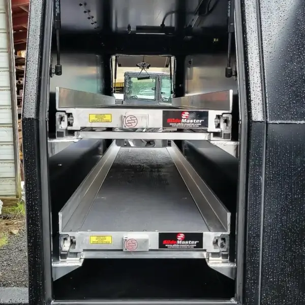 The Wildcat 1150 features two empty, extendable SlideMaster metal storage drawers for wildfire equipment inside a vehicle. Raindrops speckle the exterior, with a building and vehicle in the background—ideal for firefighter gear.