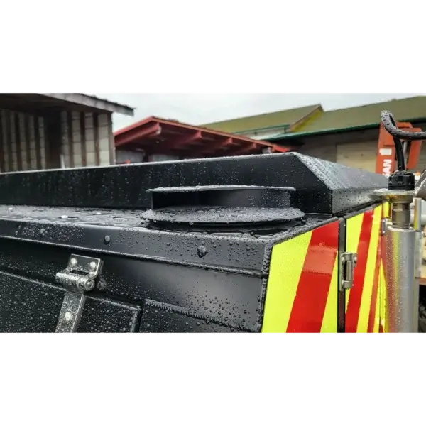 Close-up of the Wildcat 1150 wildfire equipment in Canada: a black metal unit with water droplets, circular top vent, and red/yellow reflective stripes on the side; sheds and buildings are seen in the background.