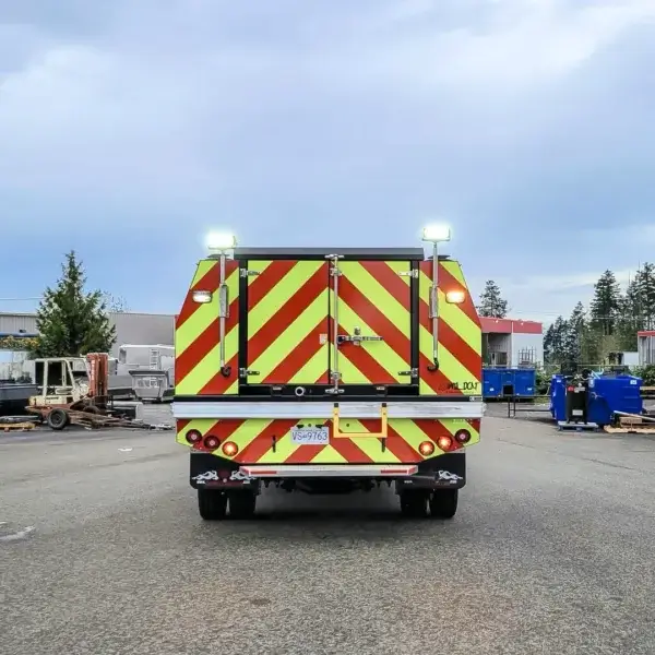 A Wildcat 950 utility truck with bright yellow and red diagonal stripes, commonly used for wildfire response in Canada, is parked on an industrial lot surrounded by buildings, equipment, and trees.