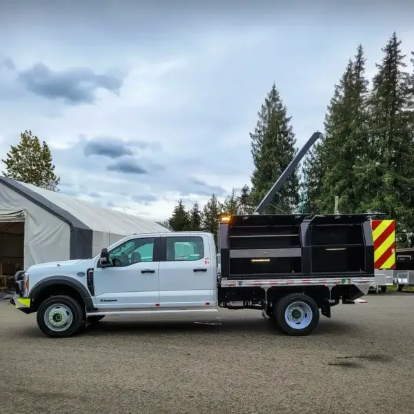 A Wildcat 950 white flatbed truck with wildfire equipment, a black industrial container, and a red-and-yellow striped rear is parked on asphalt near a large white tent and tall evergreen trees under a cloudy sky.