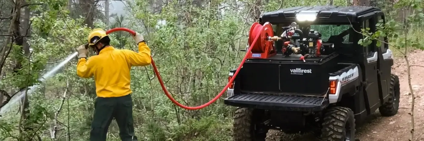 A firefighter in wildfire gear uses a hose attached to a specialized off-road vehicle to spray water on trees in a forest, working to control or prevent a wildfire.
